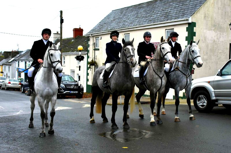 Four grey horses with riders in hunt attire, standing in the middle of a street